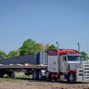 This image, provided by Steel Concepts, showcases the transportation of pre-fabricated steel structures. A prominent Peterbilt semi-truck, featuring a red and white cab, is parked with a flatbed trailer securely loaded with substantial dark gray steel components. This scene effectively illustrates the logistical capabilities for delivering large-scale steel elements to construction sites, vital for efficient project execution.