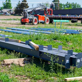 This image, provided by Steel Concepts, depicts steel structural components prepared for construction on a greenfield site. Several large, dark grey steel beams are arranged in the foreground, indicating initial stages of building development. In the background, a telehandler and a flatbed truck loaded with additional steel materials are visible, underscoring the logistical aspects of steel construction projects.