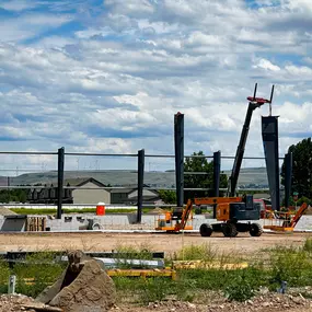 This image captures a critical phase of steel building construction by Steel Concepts, highlighting the precise erection of steel columns. A large boom lift or crane is actively positioning a column, demonstrating the sophisticated equipment and skilled labor involved in creating robust structural frameworks. The partially completed structure stands on a prepared foundation, with additional steel beams awaiting installation. In the background, residential buildings and wind turbines suggest the 