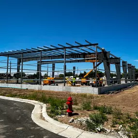 This image prominently features the ongoing erection of a steel frame building, expertly constructed by Steel Concepts. The clear blue sky provides a strong backdrop for the precisely aligned structural steel components, including columns, beams, and roof purlins, which define the skeleton of the future facility. Construction equipment and workers are visible within the frame, actively engaged in the assembly process, underscoring the efficiency and precision of Steel Concepts' operations.