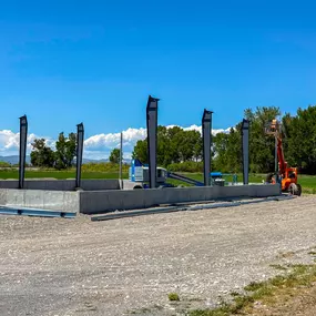 An outdoor image showcasing a foundation installation by Steel Concepts Inc. The image features a rectangular concrete foundation with several dark grey steel columns already erected and secured. The surrounding area is a gravel construction site, with green fields and trees in the background under a clear blue sky. A blue boom lift and parts of an orange lift are visible in the distance, suggesting ongoing work or readiness for further construction. This image captures an early stage of a struc