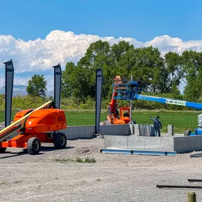 An outdoor image depicting a construction site managed by Steel Concepts Inc., showcasing the initial stages of a metal building project. The scene features a concrete foundation with several dark grey steel columns already in place, forming part of the structure's framework. Heavy machinery, including an orange boom lift and a blue 