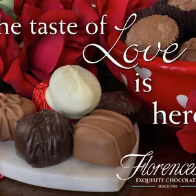 A Valentine's Day-themed table featuring Florence's Exquisite Chocolates in Rexburg, Idaho. The table is adorned with a variety of chocolate truffles displayed on a white marble heart-shaped plate, surrounded by red roses and a red bowl with white hearts containing more chocolates. Text on the image reads, 