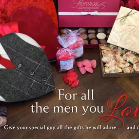 A Valentine's Day-themed display featuring Florence's Exquisite Chocolates in Rexburg, Idaho. The setup includes a variety of beautifully packaged chocolates and confections, such as a heart-shaped box designed to look like a suit with a red tie, a gold box filled with nut brittle, and an assortment of truffles and candies. The display is adorned with red roses and heart-shaped decorations, creating a romantic and inviting atmosphere. The text on the image reads, 