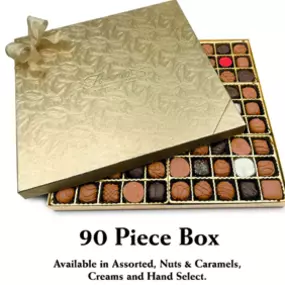 This photograph features a 90-piece box of Florence's Exquisite Chocolates, likely from their Rexburg, Idaho location. The chocolates are packaged in a luxurious gold box, tied with a large, elegant bow. The lid of the box is partially open, revealing a meticulously arranged assortment of chocolates. The chocolates vary in type, including nut clusters, caramels, creams, and other specialty pieces.