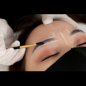 Woman getting microblading, 2 hands with white gloves, and esthetician markeing area of eyebrows. Black mask.