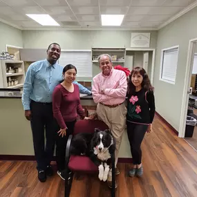 A group of four smiling staff members and a friendly black and white dog in the reception area of the Chiropractic Wellness Group office. The image conveys a warm and welcoming atmosphere.