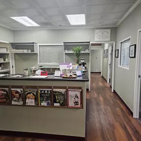 A clean and welcoming chiropractic office reception area, featuring a front desk, a magazine rack for patients, and a hallway leading to treatment rooms. The space has wood-style flooring and light green walls, creating a professional and organized environment.