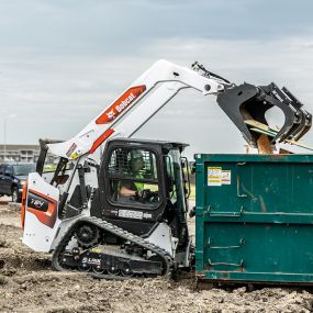 Construction Worker Using Bobcat T64 Track Loader With Industrial Grapple Attachment To Drop Plywood Into Jobsite Dumpster