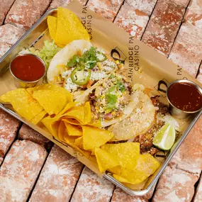 A mouthwatering platter of tacos and chips with salsa served at the sportsbook restaurant and bar at Plainridge Park Casino in Plainville, Massachusetts. There are three tacos - one beef birria taco topped with beef, pickled onions, lettuce, cilantro and cheese; one pork carnitas taco with pork, corn salsa and cilantro; and one chicken taco with jalapeno, pineapple, crema and cilantro. The tacos are accompanied by fresh crunchy corn tortilla chips and a side of salsa. The combination of tacos an