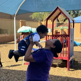 A caring teacher lifting a child up with encouragement and joy, celebrating the fun and excitement of playtime.