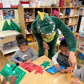 Learning through fun and imagination! The teacher dressed as a dinosaur adds a playful twist while helping with art projects.