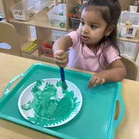 A girl concentrates on adding green paint to her artwork, blending colors on the page.
