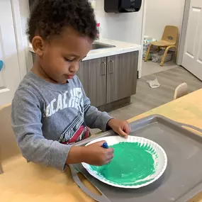 A boy dips his brush into green paint, creating shapes and patterns on white paper.