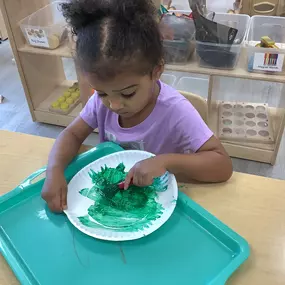 A little girl smiles as she uses green paint to decorate her artwork at a classroom table.
