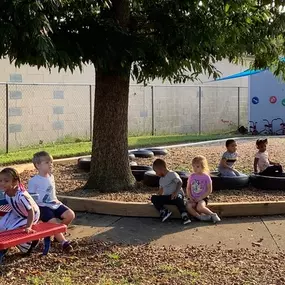 A group of children gathered together under a tree, engaged in quiet play and enjoying the natural surroundings.

Alt text: