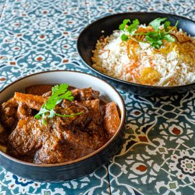 Bowl of Lamb Vindaloo in a blue serving bowl, aesthetically framed by a bowl of Jeera Rice and garnished with cilantro, placed on a blue tiled surface