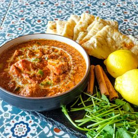 Bowl of Murgh Lababdar in a blue serving bowl, aesthetically framed by cilantro, whole lemons, naan cut into quarter pieces, and cinnamon sticks, placed on a colorful black and white tiled surface