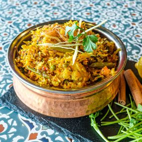 Bowl of Vegetable Biryani in a hammered copper serving bowl, aesthetically framed by cilantro, whole lemons, and cinnamon sticks, placed on a colorful blue tiled surface