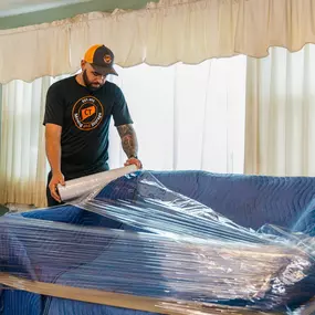 A mover wraps a sofa with moving blankets and stretch wrap for protection.