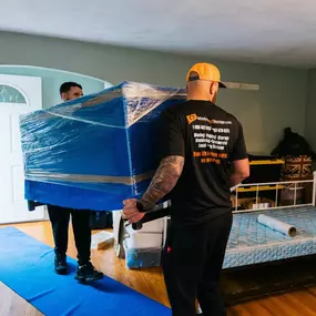 Movers carry a large, wrapped piece of furniture on a protective floor runner.