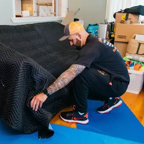 A mover places a black quilted furniture blanket over a couch to prepare it for wrapping.