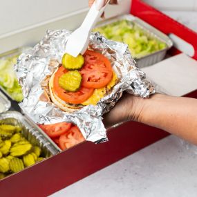A close-up photo of a person putting pickles on their cheeseburger in front of a Five Guys catering box of toppings.
