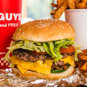 A close-up photograph of a Five Guys cheeseburger, soft drink in red Five Guys cup, and regular order of fries sitting on a table inside a Five Guys restaurant.
