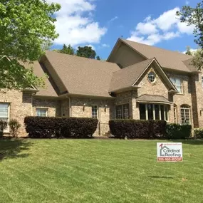 Light brown-colored brick home with a brown asphalt shingle roof displaying a Cardinal Roofing sign in the front yard