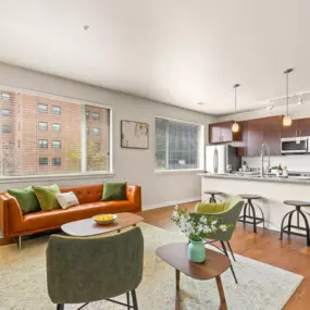 Modern living room with an orange couch, green accent chairs and oversized windows, complete with view into kitchen with barstool seating and pendant lighting