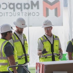 A group of construction workers from Mappco Construction are gathered on a work site, engaged in a detailed discussion. They are all wearing white safety helmets and high-visibility vests with the Mappco logo. The setting suggests an active construction environment where teamwork and planning are underway.