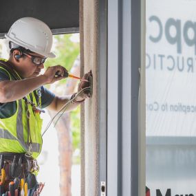 A construction worker wearing a white helmet and high-visibility vest is installing electrical wiring in a wall socket. The worker is using a screwdriver and is equipped with a tool belt full of various tools and cables.