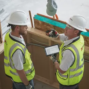 Two Mappco Construction professionals are in the midst of a project site consultation. Chad Mapp, on the right, is holding a tablet, displaying blueprints or project plans, while gesturing towards an area of interest. They stand within a partially completed structure, surrounded by building materials and insulation layers.