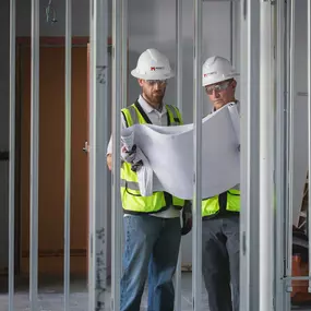 Experienced Mappco Construction professionals meticulously review building plans on-site. Two diligent construction managers stand in high-visibility vests and safety helmets engrossed in project blueprints amidst the skeletal framework of metal studs, highlighting Mappco's hands-on approach to ensuring precision and quality in commercial construction projects.