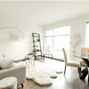 A light filled living room with large windows, a glass sliding door to the patio, filled with a white couch, white rug, and a small table of chairs