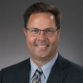 Professional headshot of John Burns, CEO of Exencial Wealth Advisors, wearing a suit and striped tie with a grey background.