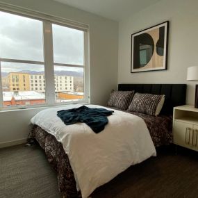 A cozy bedroom with oversized windows that view the foothills of Boise with a bed, mightsand with gold hardware, and desk lamp