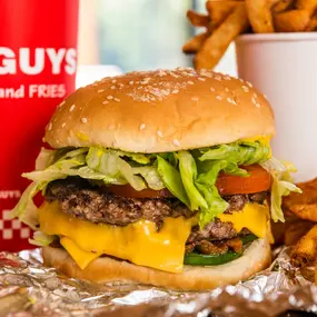 A close-up photograph of a Five Guys cheeseburger, soft drink in red Five Guys cup, and regular order of fries sitting on a table inside a Five Guys restaurant.