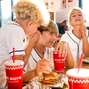 Children chat and laugh while enjoying their Five Guys meals at a dining room table in a Five Guys restaurant.