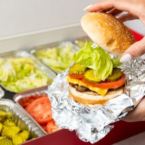 A close-up photo of a person putting the top bun of their cheeseburger in front of a Five Guys catering box of toppings.