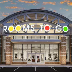 Rooms To Go store exterior at dusk with illuminated logo and modern glass-front design.