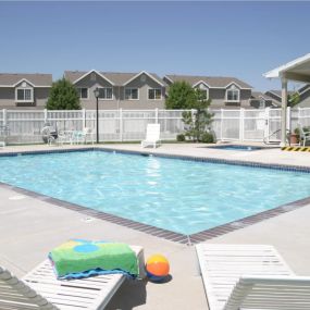 View of the blue swimming pool with a white fence, white chaise lounge chairs with a colorful towel on one and a multicolored beach ball in between, and resident building to the right