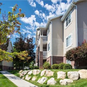 Side view of Country Oaks Apartment building with columns featuring part brick work, large oversized windows, white balcony railings, and manicured pathway with rock landscaped yard area