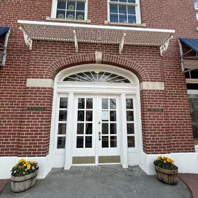 A close-up of a building entrance with a red brick facade, a central arched doorway with white double doors, and a decorative white metal canopy above it.