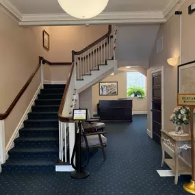 An interior lobby with a dark blue patterned carpet, light pink walls, a large staircase with dark wood handrails, and a view into a small reception area under the stairs.