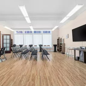 A bright meeting or event room with light wooden floors, pink walls, rows of black folding chairs, and two large, black screens mounted on the right wall.