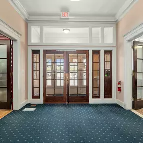 The lobby entrance of a building with a dark blue patterned carpet, light pink walls, and central glass-paneled double wooden doors flanked by open side doorways and a red EXIT sign above.