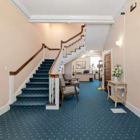 An interior hallway or lobby with light pink walls, a dark blue patterned carpet, and a white staircase with dark wood handrails leading up and turning right.