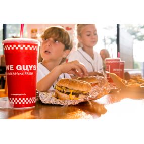 A red Five Guys soft drink cup is the focus in the foreground, as children enjoy their meals in the background.