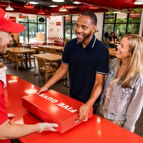 A couple collect their Five Guys catering box from a Five Guys employee at a Five Guys restaurant.